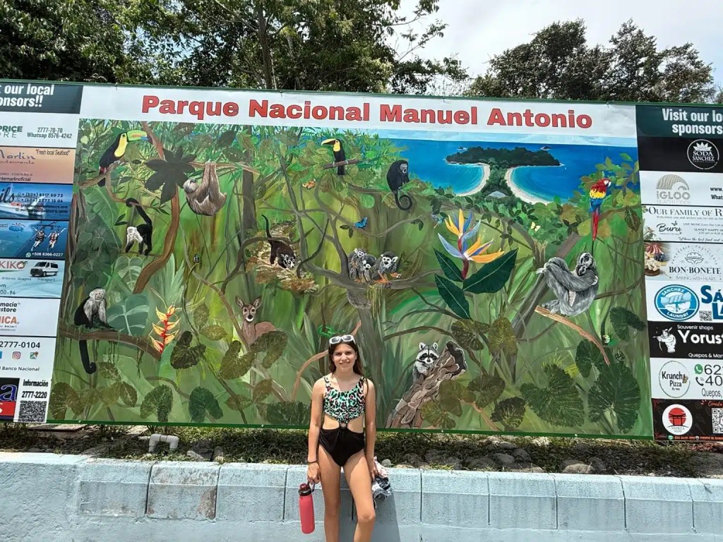 A girl in a swimsuit stands beside a colorful mural depicting wildlife and scenery from Parque Nacional Manuel Antonio, featuring various animals and a tropical landscape.
