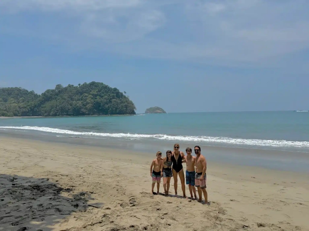 A group of five people standing on Manuel Antonio beach with gentle waves in the background and a lush green hillside in the distance. They appear to be enjoying a sunny day at the beach.