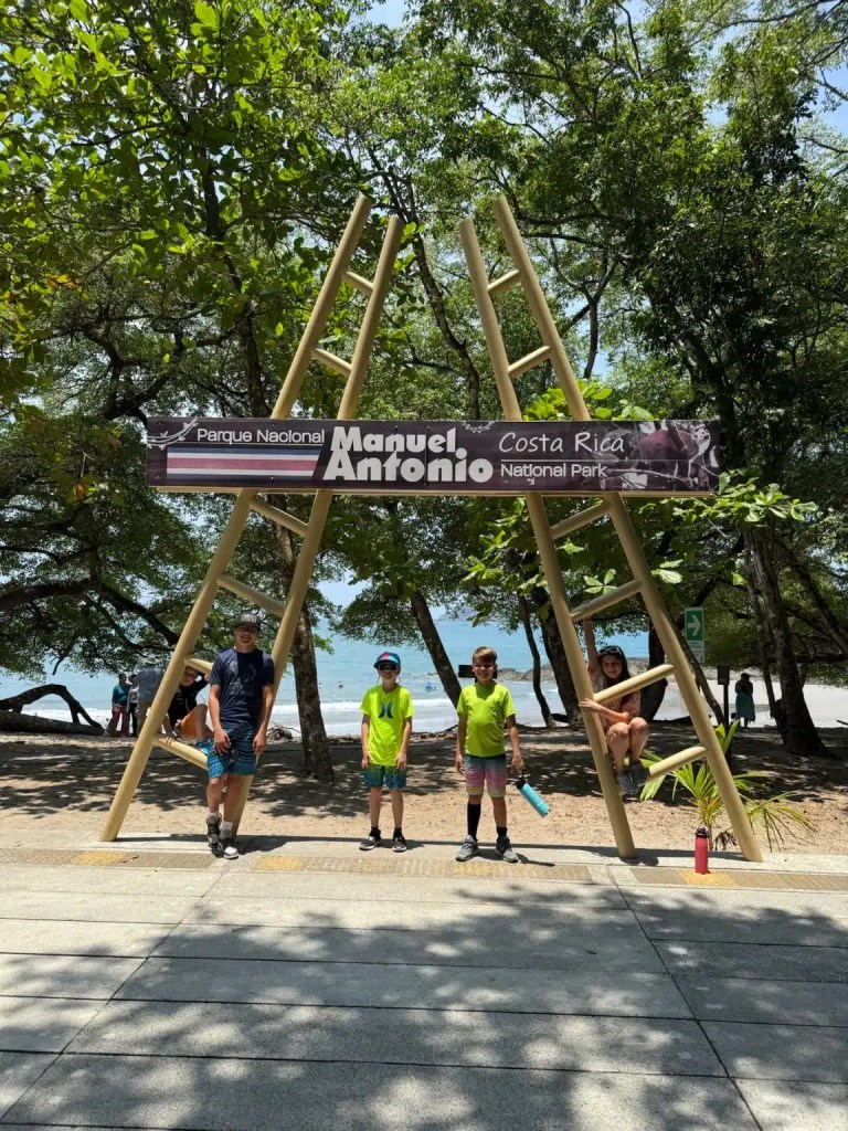 Children posing in front of the Manuel Antonio National Park sign in Costa Rica, surrounded by trees and beach in the background.