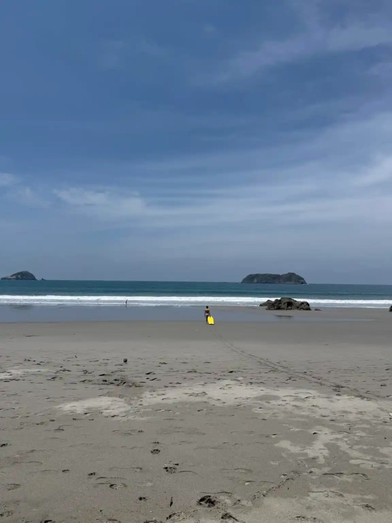 A sandy Manuel Antonio beach with gentle waves and a clear blue sky, featuring a person with a yellow surfboard walking towards the water.