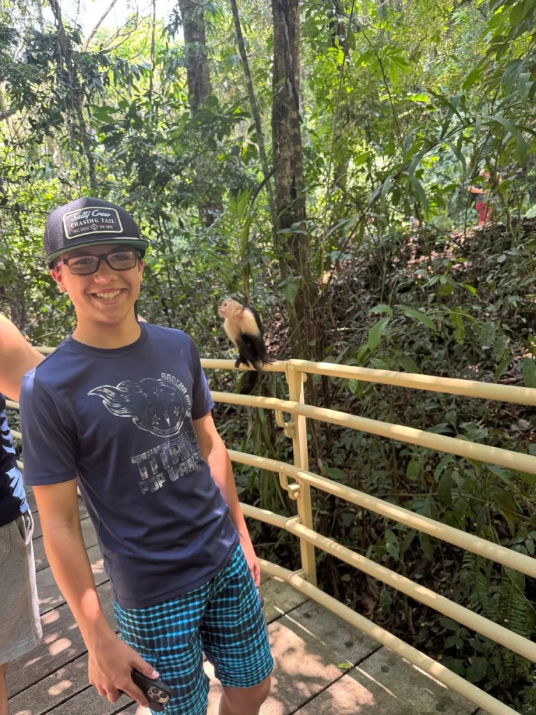 A young person smiling for the camera while standing on a path in a lush green jungle, with a monkey visible in the background.
