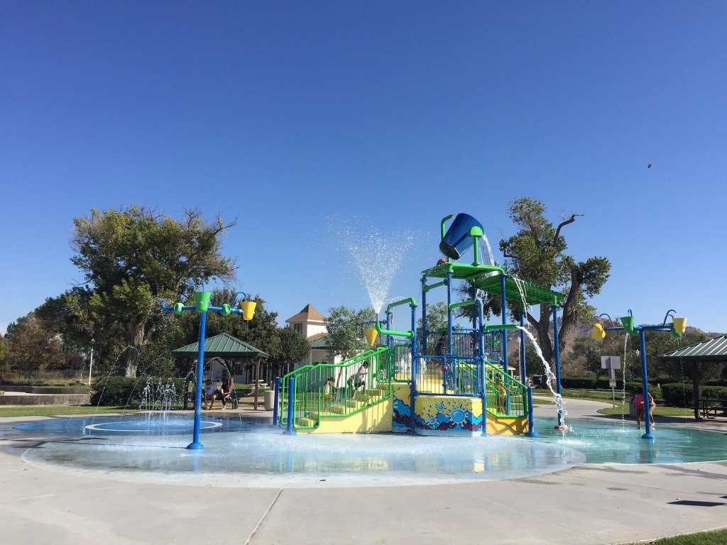 A colorful water play structure with slides and water features in a park, surrounded by trees and clear blue skies.