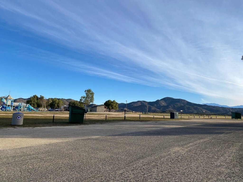 A park scene featuring a playground in the background, green hills, and a clear blue sky. Several trash bins are visible along a gravel pathway at this Riverside campground.