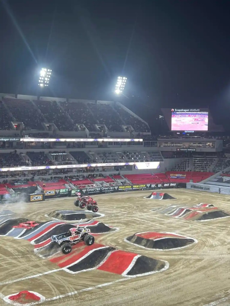 View of the Monster Jam event at Snapdragon Stadium, featuring monster trucks on a dirt track with stadium lights illuminating the scene and a crowd in the background.