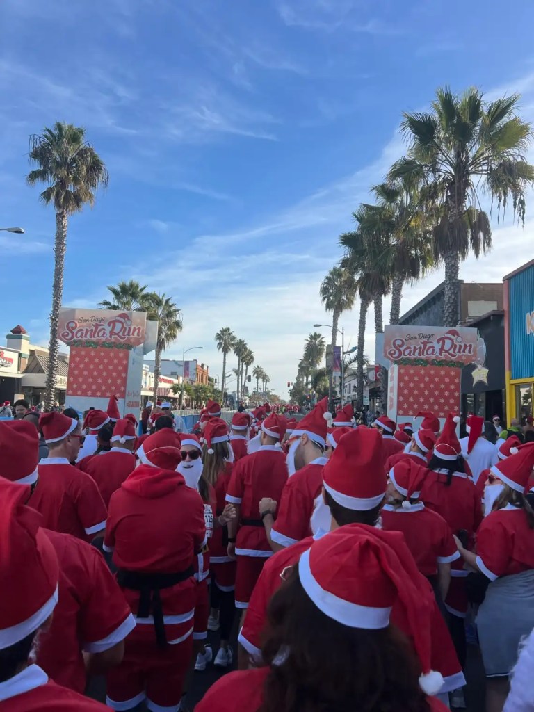 A crowd of participants in Santa suits with festive hats gathered for the San Diego Santa Run, lined up along a palm tree-lined street under a clear blue sky.