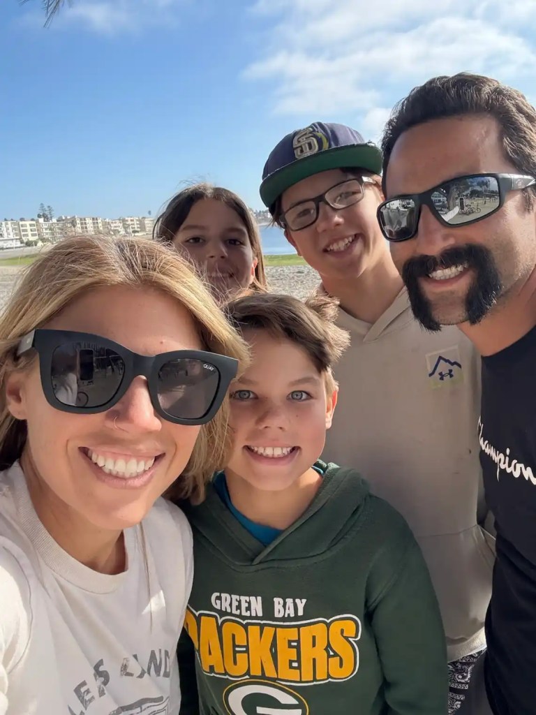 A group selfie at a San Diego beach in winter featuring five smiling individuals, with sunlight and a picturesque coastal background.