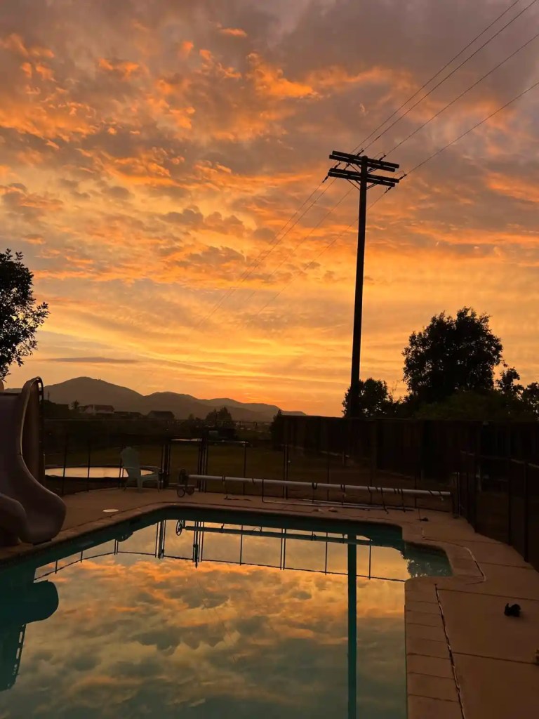 A vibrant winter sunset scene in San Diego with orange and pink hues reflecting in a calm swimming pool, surrounded by a grassy area and silhouetted trees, with mountains in the background.