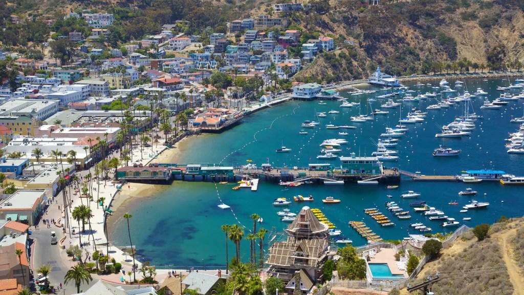 Aerial view of a vibrant coastal town with colorful buildings, a marina filled with yachts, and sandy beaches under a clear blue sky.