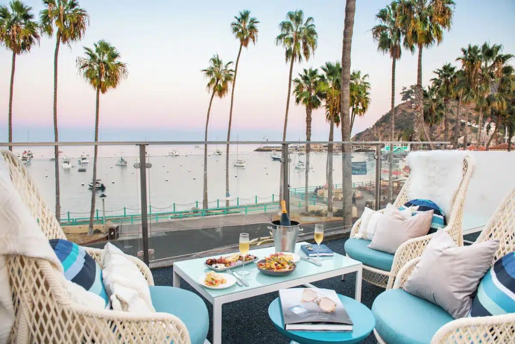 A cozy outdoor seating area with wicker chairs and a small table, offering a beautiful view of the ocean, boats, and palm trees at sunset.