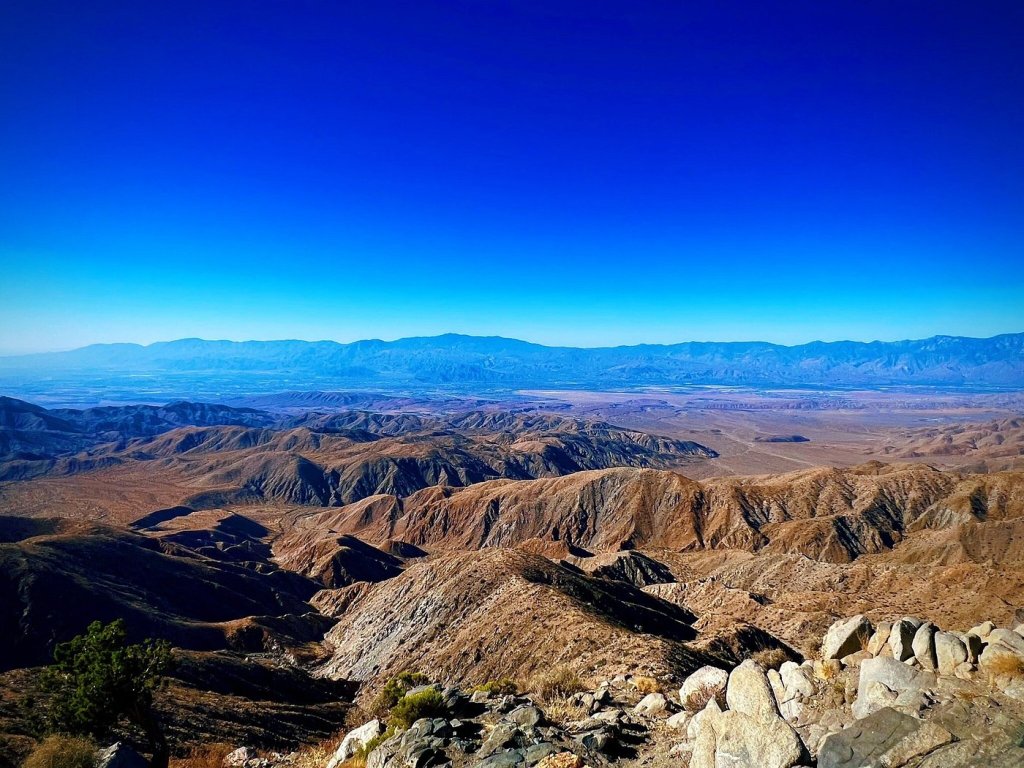 A panoramic Joshua Tree view of rugged mountains and desert terrain under a clear blue sky, showcasing layers of brown and beige hills with distant mountains on the horizon.