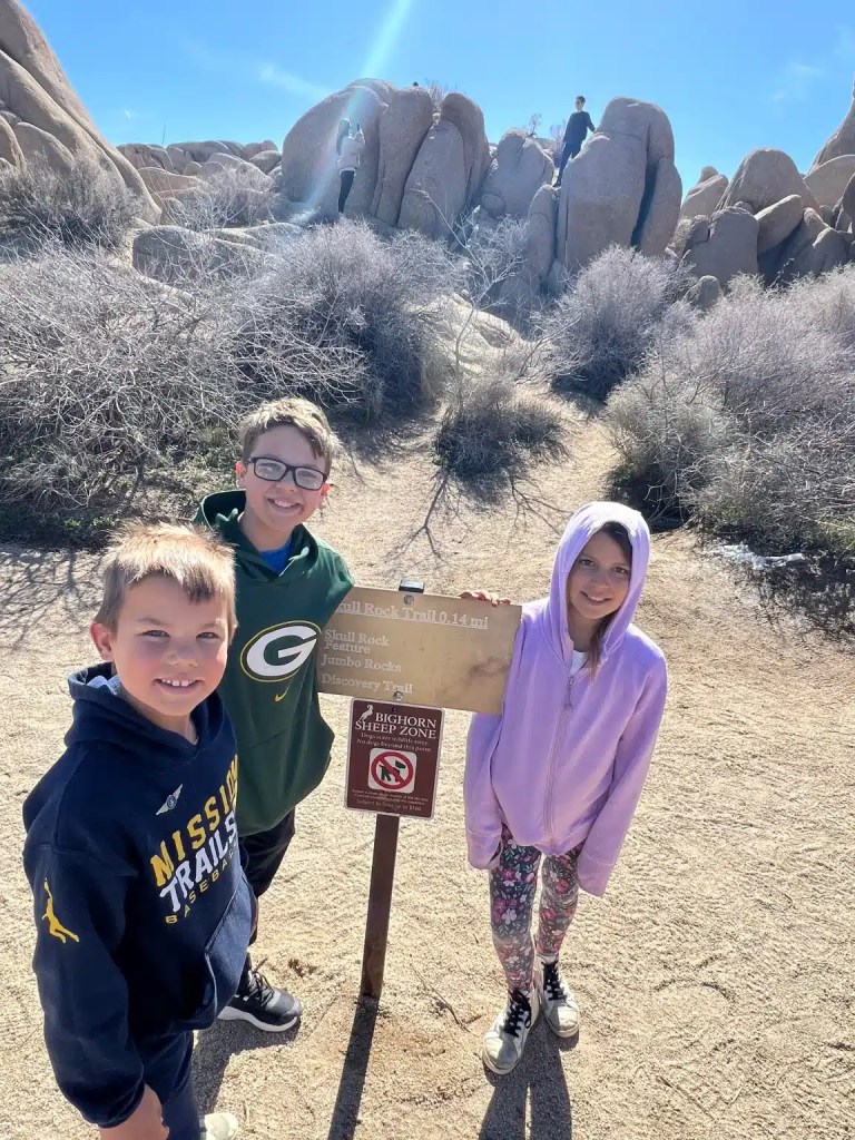 Three children smiling in front of a Joshua Tree trail sign at a rocky outdoor location, with a person climbing rocks in the background.