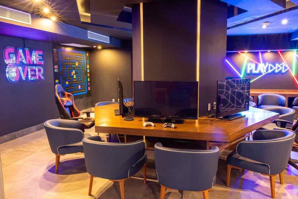 A modern gaming lounge featuring a large wooden table surrounded by blue chairs, two gaming monitors, and neon signs that read 'GAME OVER' and 'PLAY' at one of the best all inclusive resorts for teens.