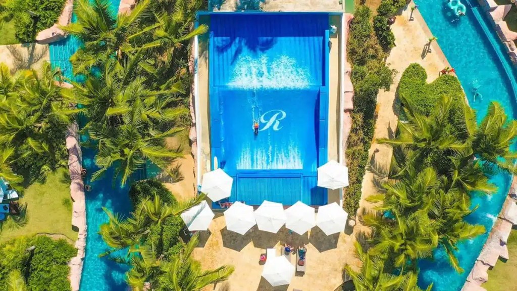Aerial view of a blue water park area at a teen-friendly all inclusive resort featuring a wave pool, surrounded by palm trees and lounge chairs with umbrellas.