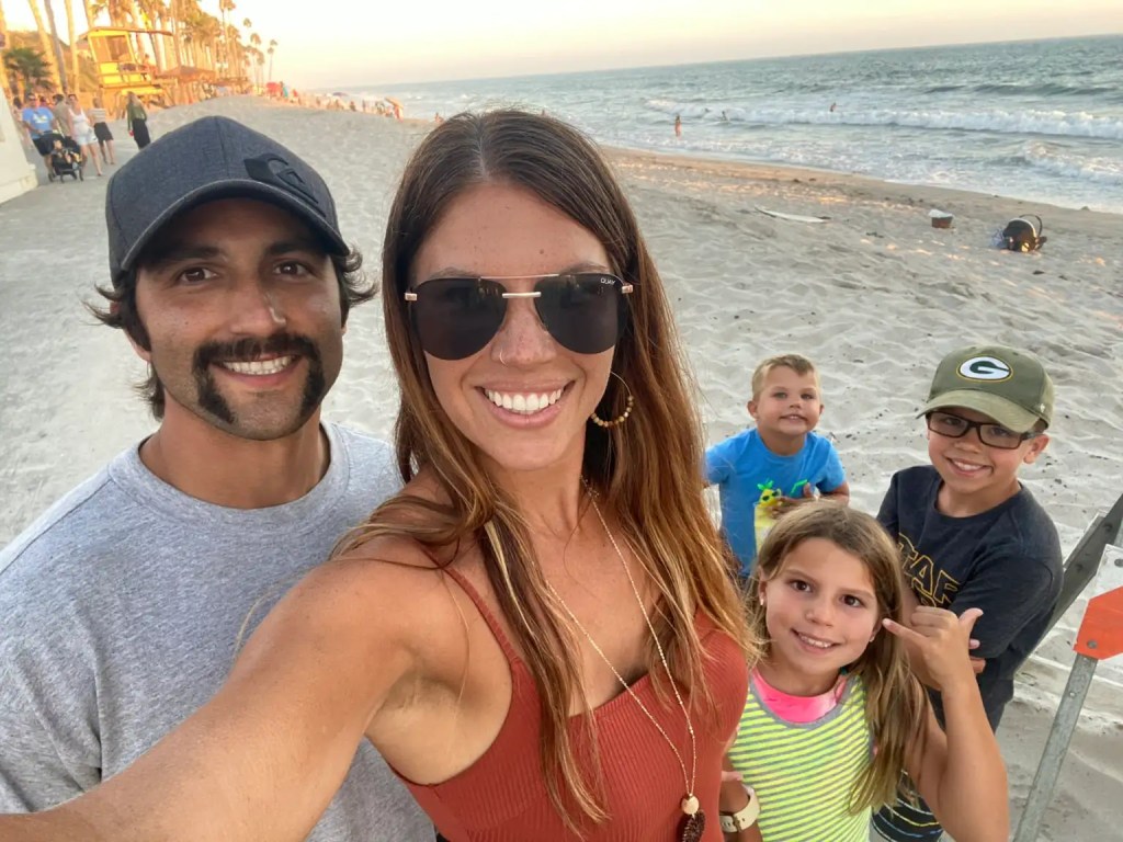 A family poses for a selfie on one of the best beaches in CA during sunset, with palm trees and the ocean in the background. The group consists of two adults and three children, all smiling and enjoying their time together.
