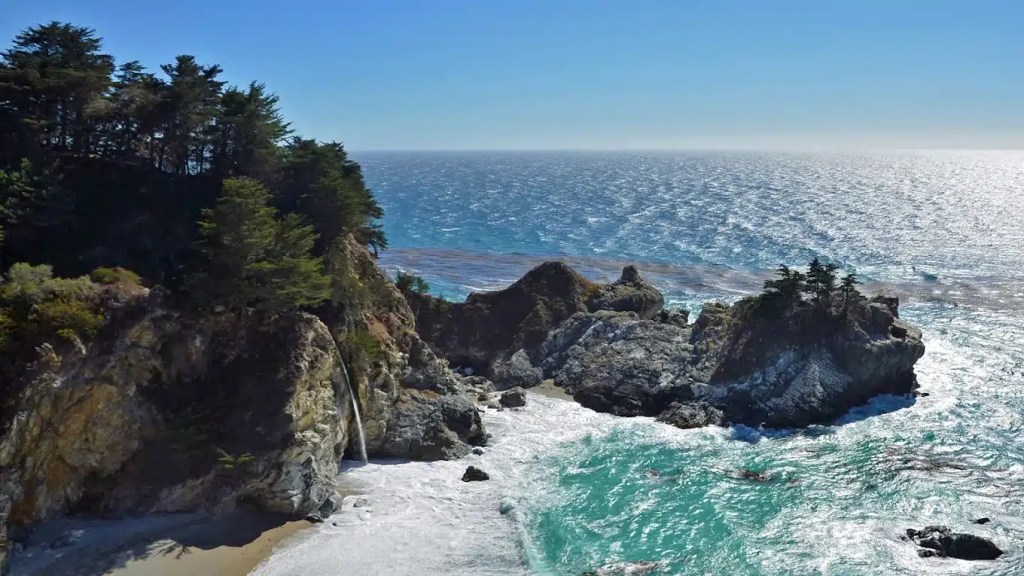 A scenic view of a rocky coastal landscape at one of the best ca beaches, with a waterfall flowing into the ocean, surrounded by lush greenery and a sparkling sea under a clear blue sky.