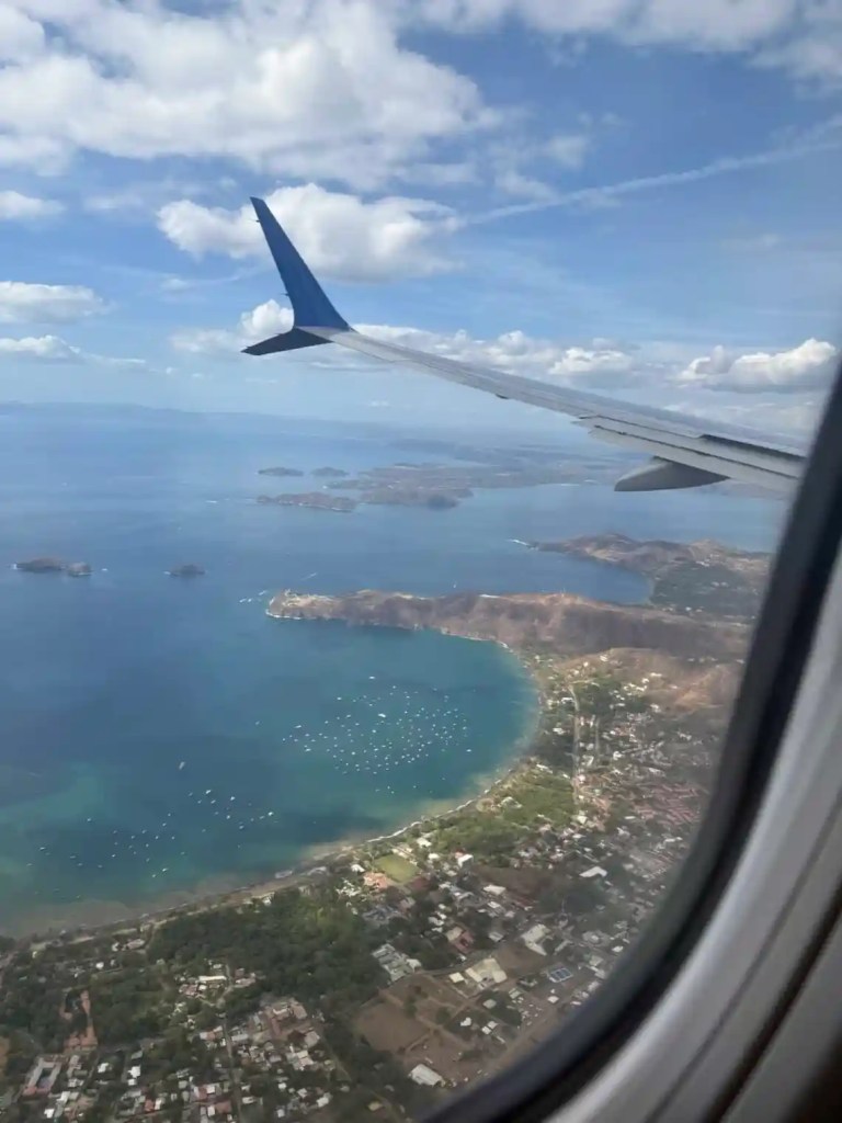 View from an airplane window showing a wing, coastline, and blue ocean with scattered boats and islands in the distance under a partly cloudy sky. Traveling to one of the countries that is warm in December.