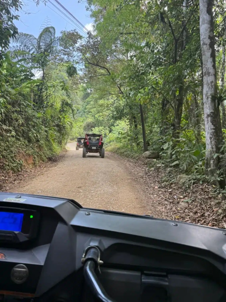 View from the front of an off-road vehicle traveling down a narrow dirt road surrounded by lush greenery and trees, with another vehicle visible ahead.
