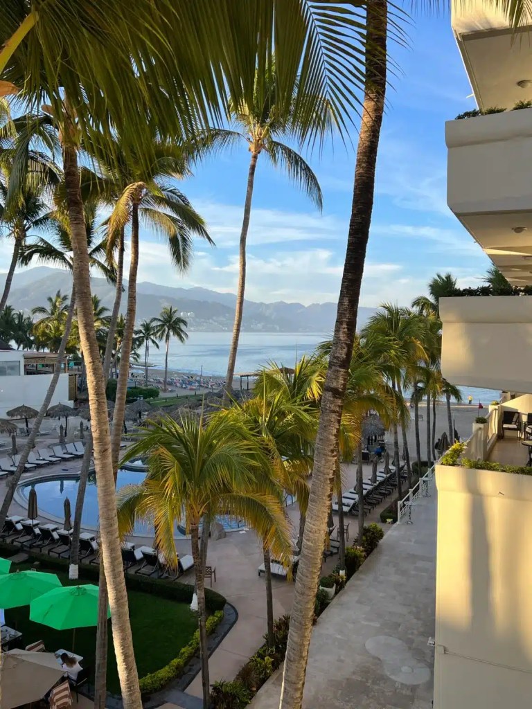 View of a tropical resort with palm trees, a swimming pool, and the ocean in the background, under a blue sky. Mexico is one of the countries that are warm in December.
