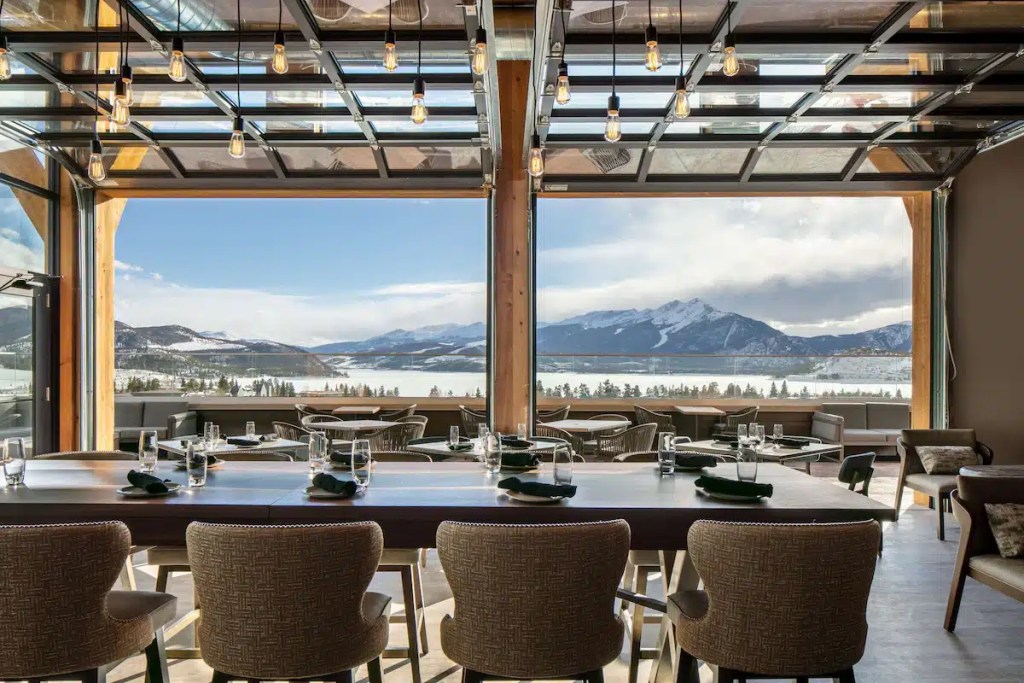 Interior view of a modern restaurant featuring a large window overlooking a snowy mountain landscape and a lake. The dining area includes a long table set with glassware and green napkins, with stylistic light fixtures hanging from the ceiling.