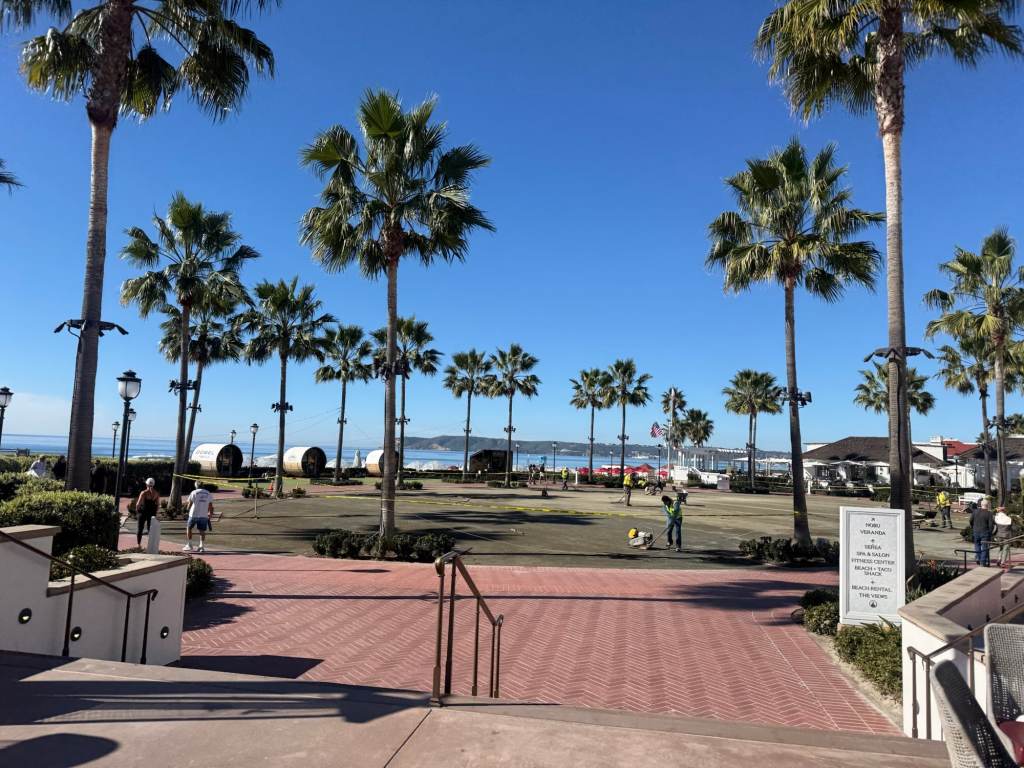 A sunny beachfront park with palm trees, brick pathways, and people walking. There are tents in the background and activities taking place, with a clear blue sky above.
