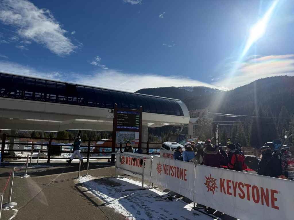 The River Run Village gondola at Keystone Resort with people waiting in line, snow on the ground, and clear blue skies in the background.