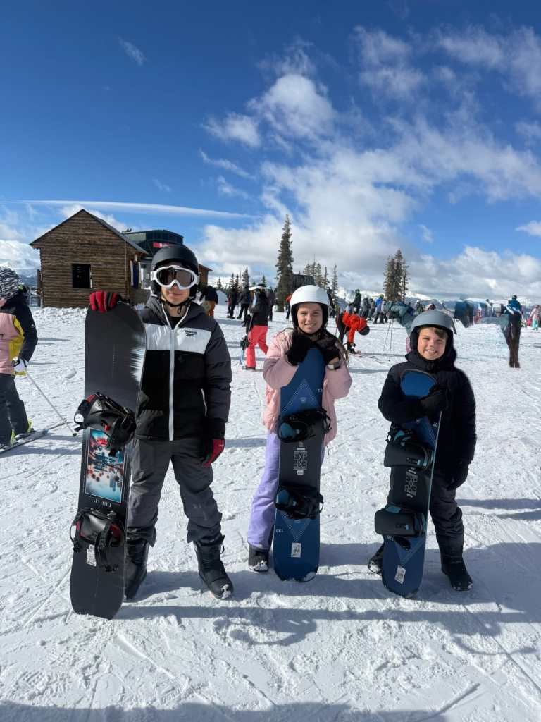 Three children with snowboards pose on a snowy slope with a wooden cabin in the background and a mountain ski resort atmosphere.