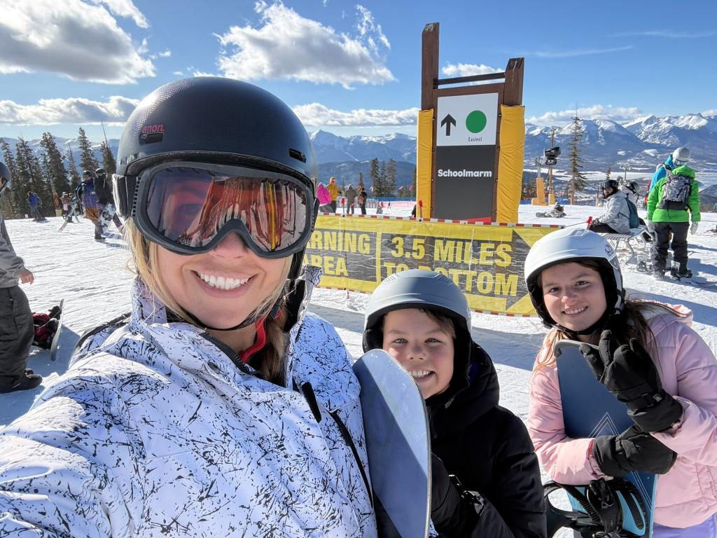 Three people smiling for a selfie on a ski slope, wearing helmets and snow gear, with a sign in the background indicating the Schoolmarm ski run and a warning about the distance to the bottom.