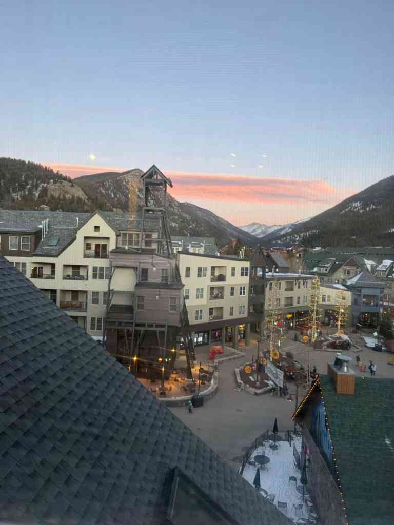 A scenic view of a River Run Village at dusk, featuring modern buildings, outdoor seating, and decorated trees, with mountains in the background and a colorful sky.