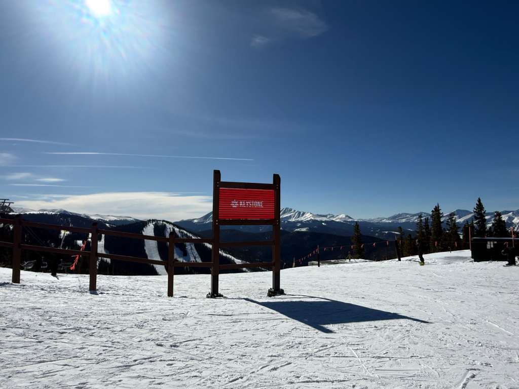 Snow-covered ski slope with a wooden sign and mountain backdrop under a clear blue sky.