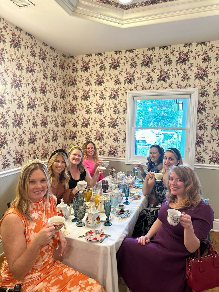 A group of eight women enjoying tea at a beautifully set table adorned with vintage tea sets, colorful glassware, and floral wallpaper.