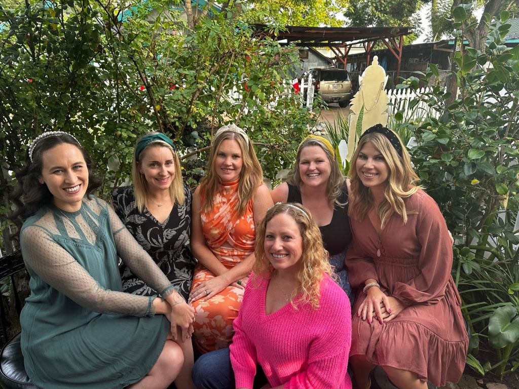 A group of six women sitting together outdoors among greenery, smiling and posing for the camera. They are dressed in various colorful outfits, with some wearing hair accessories.