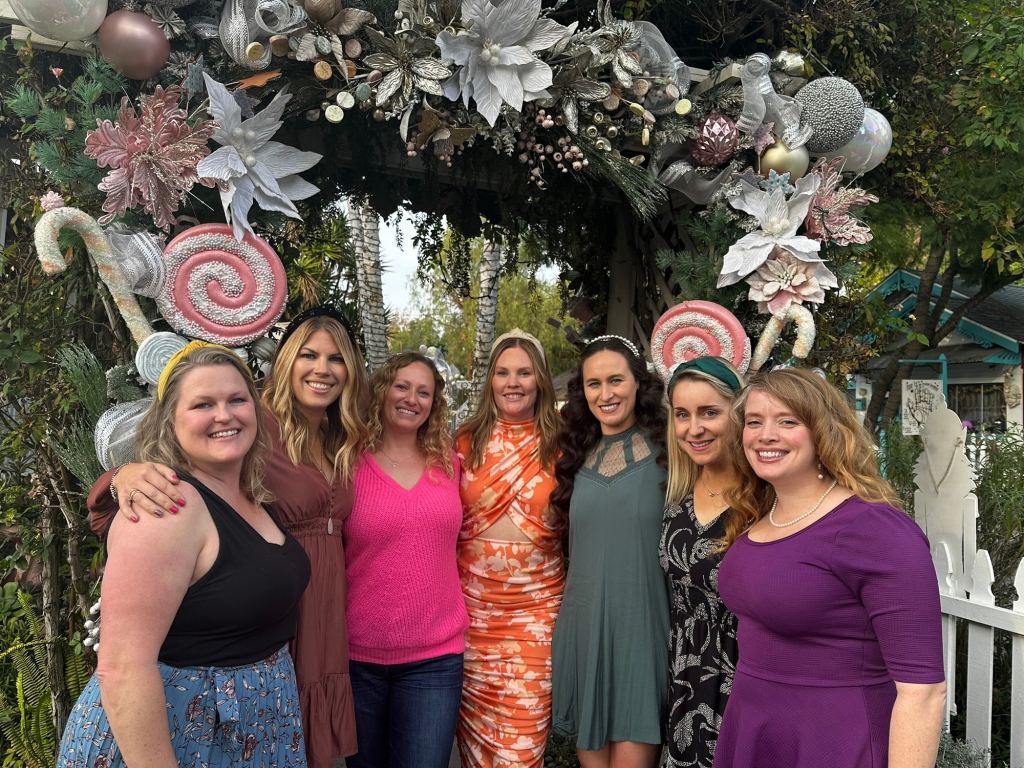 Group of seven women smiling together in front of a decorative arch at the Los Rios Tea House in San Juan Capistrano, with festive elements and greenery.