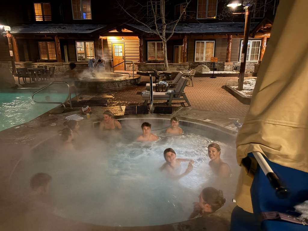 A group of people enjoying a hot tub outdoors at night in the River Run Village, with mist rising from the water. Nearby, a swimming pool is visible with lounge chairs and a wooden building in the background.