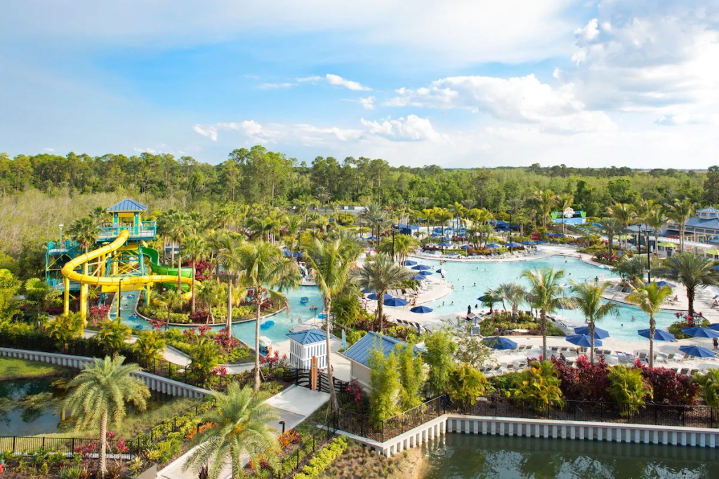 Aerial view of a vibrant water park hotel in Orlando featuring a large yellow water slide, multiple swimming pools, a lazy river, palm trees, and lounge areas with umbrellas. The background includes lush greenery and a clear blue sky.