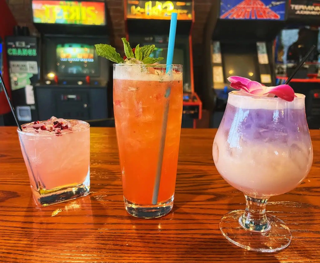Three colorful cocktails on a wooden bar with vintage arcade machines in the background. The drinks include a pink beverage garnished with dried flowers, an orange drink with a mint leaf and a blue straw, and a purple cocktail topped with a flower.