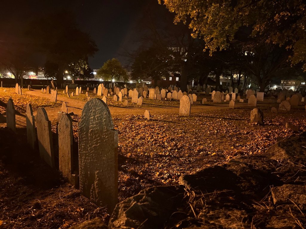 A cemetery at night in Salem, Ma, illuminated by soft orange light, featuring numerous gravestones amidst fallen leaves.