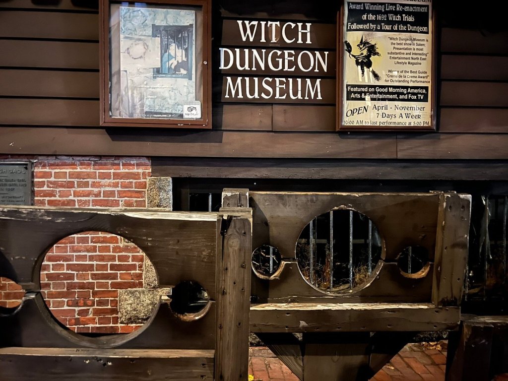 Exterior view of the Witch Dungeon Museum featuring a sign, historical artifacts, and a wooden stockade. Visiting this museum is one of the best things to do in Salem, Ma.