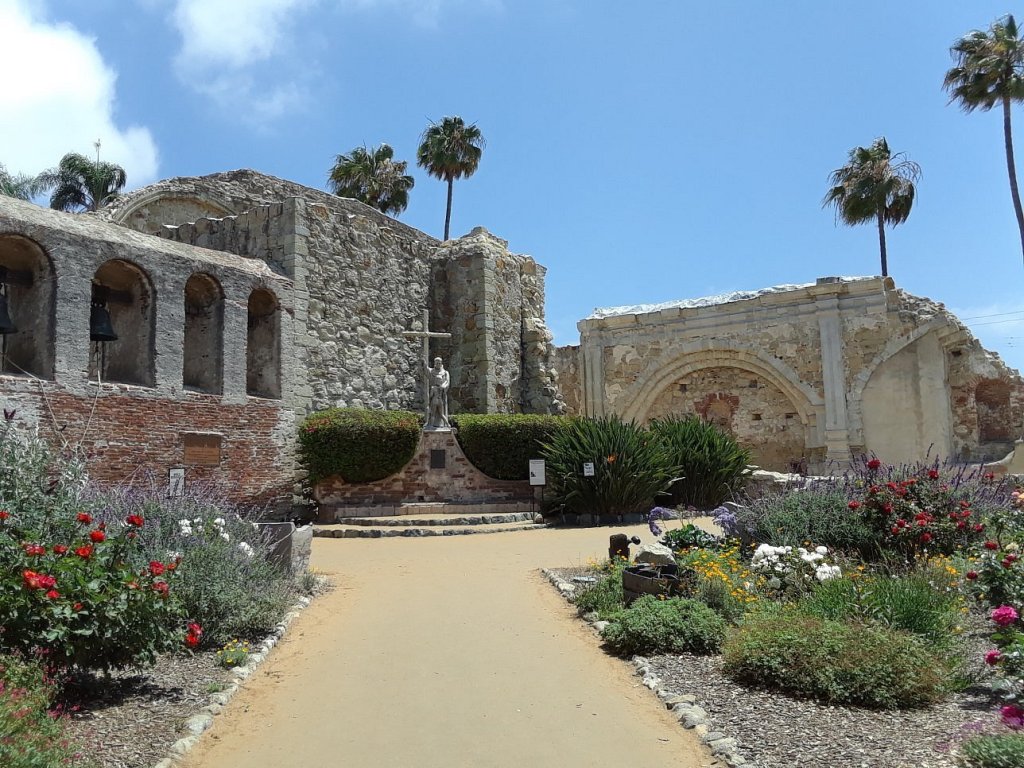 Historic ruins of the Mission San Juan Capistrano with stone walls and arches, surrounded by vibrant flower gardens and palm trees under a clear blue sky.