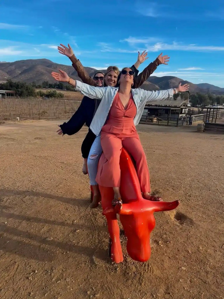 Four women posing joyfully while sitting on a red bull statue in an outdoor setting with mountains in the background at one of the Valle de Guadalupe wineries.