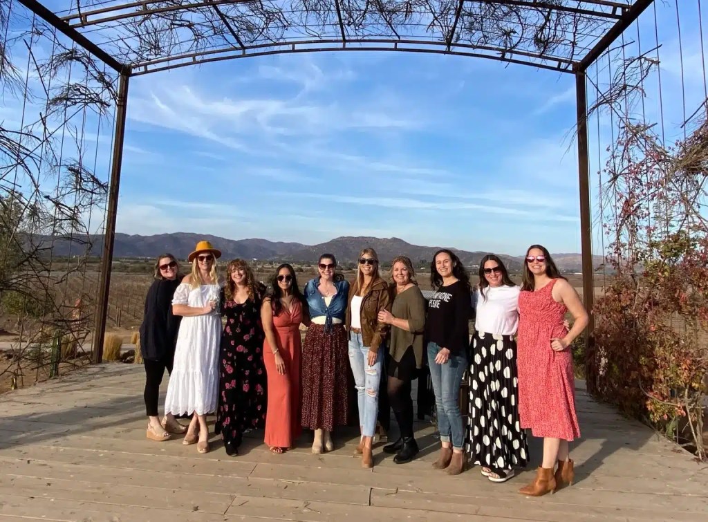 A group of ten women standing on a wooden platform at one of the Valle de Guadalupe wineries with mountains in the background, wearing various stylish outfits and smiling at the camera.