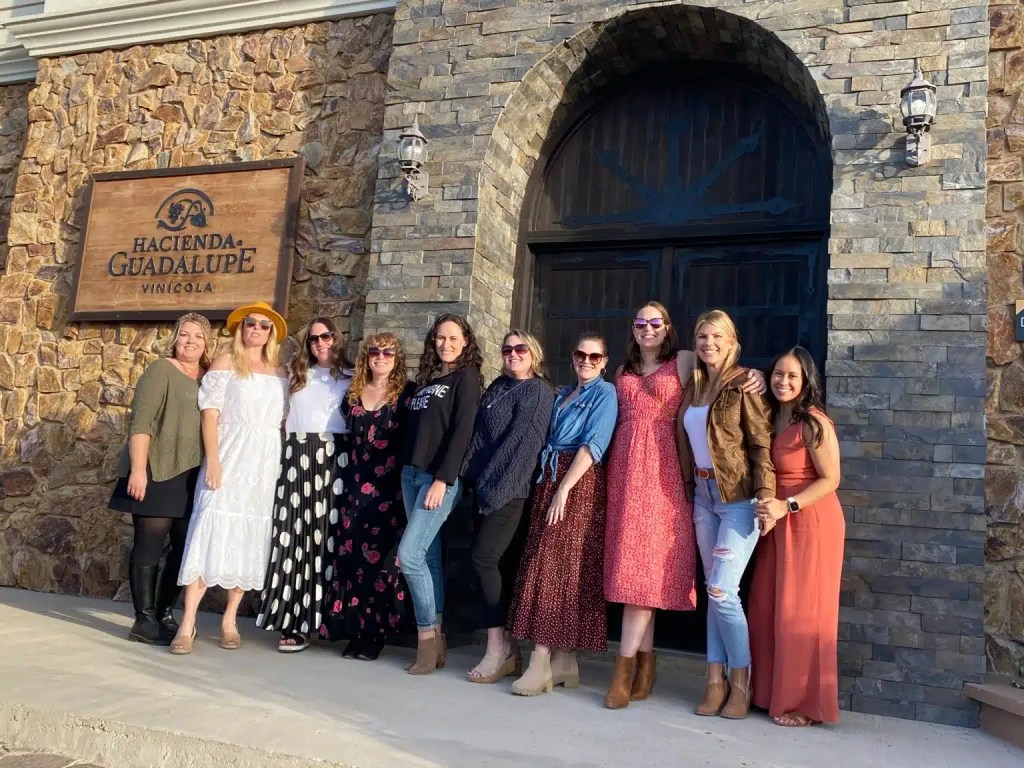 A group of twelve women posing in front of a stone building with a wooden sign that reads 'Hacienda Guadalupe Vinícola.' They are wearing a variety of colorful outfits and sunglasses, smiling for the camera.