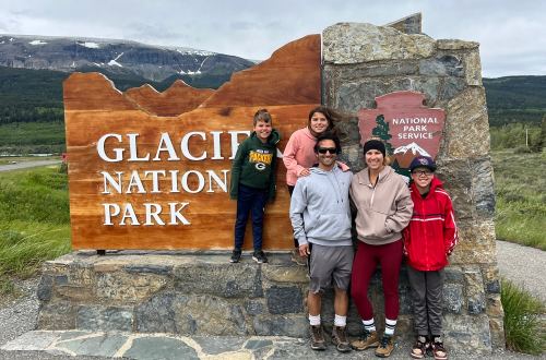 A family of five posing in front of a large sign that reads 'Glacier National Park' and 'National Park Service'.