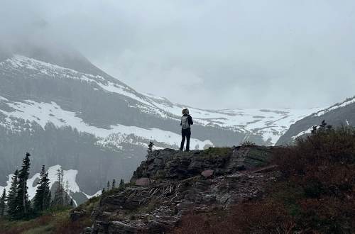 A person overlooks a snowy, mist-covered mountain landscape with views of the Grinnell Glacier from a rocky ledge.