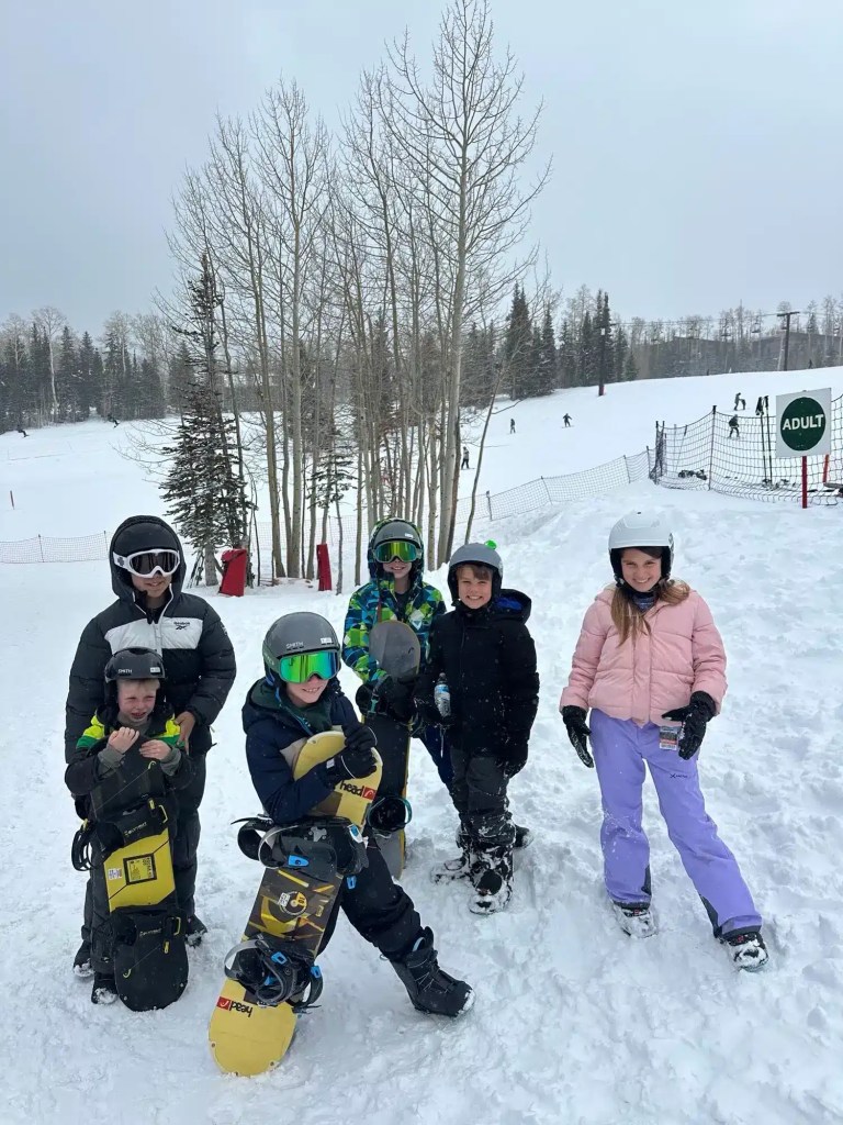 brian-head-ski-resort-hotels-10 Group of children in winter gear holding snowboards on a snowy slope near an adult ski area sign while on a family vacation in Utah.