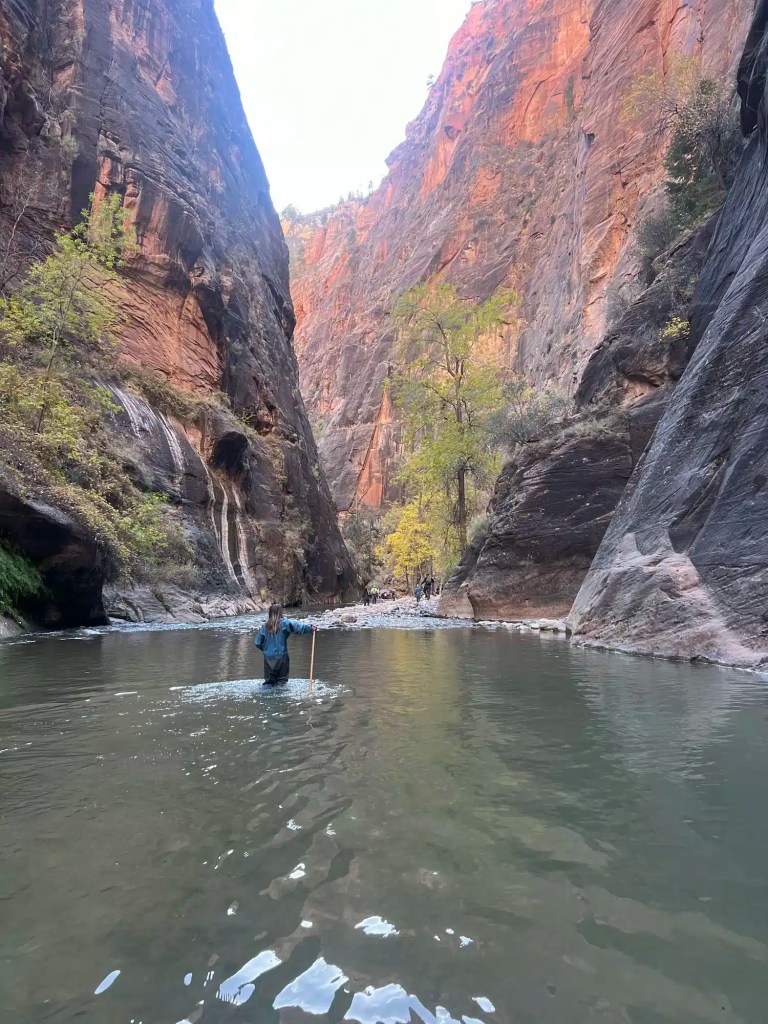 img_6508 Person crossing a shallow river in a canyon with tall red rock walls and green trees on a family vacation in Utah.