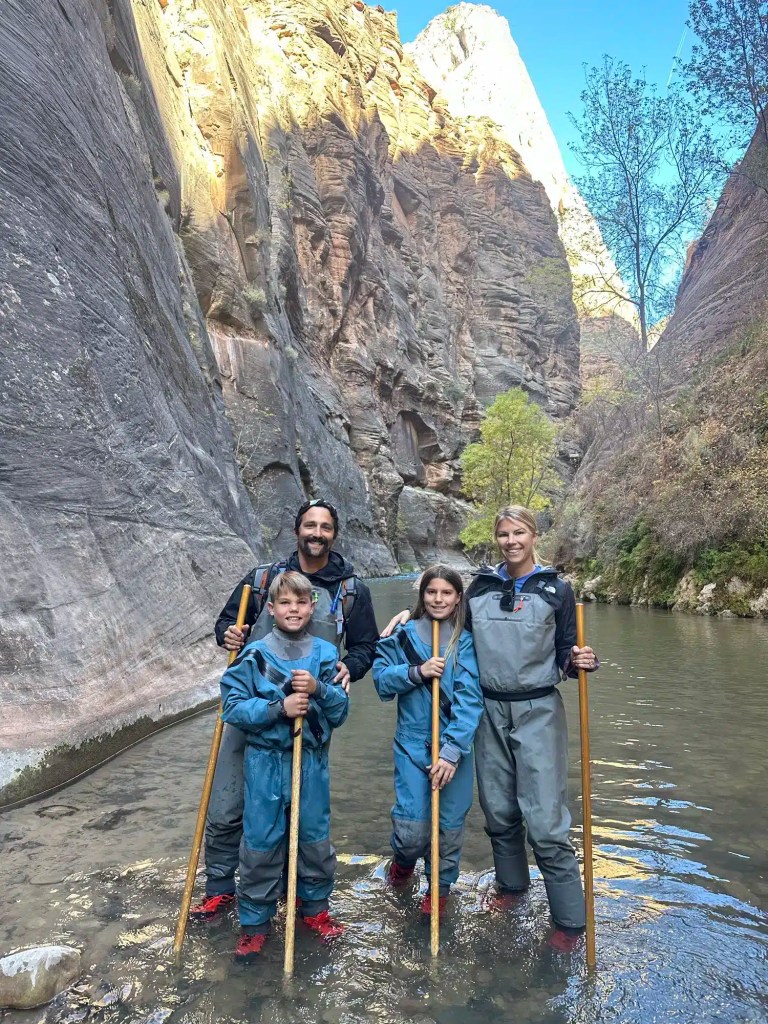 Zion-with-kids-7 A family of four standing in shallow river water within a rocky canyon, wearing waterproof wading suits and holding wooden hiking sticks while on a family vacation in Utah.