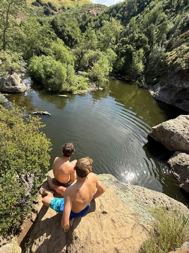 Two young men sit on a rocky ledge overlooking a sunlit pool in a canyon at Malibu Creek State Park.